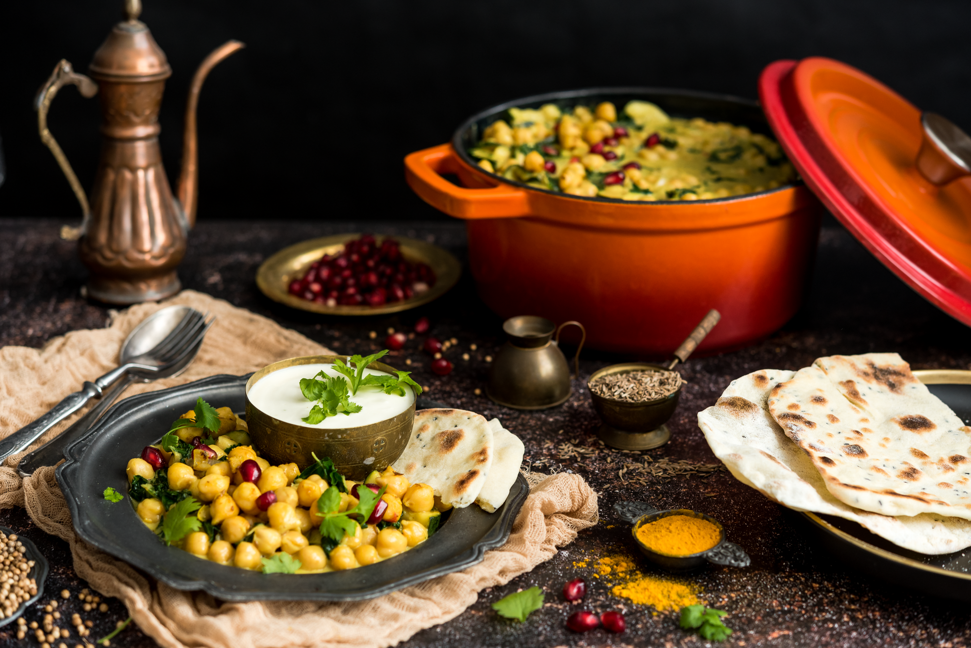 Garnek żeliwny emaliowany z potrawką z ciecierzycy. Obok garnka stoi metalowy talerz, a na nim chlebki pita, sos i danie z ciecierzycy.
