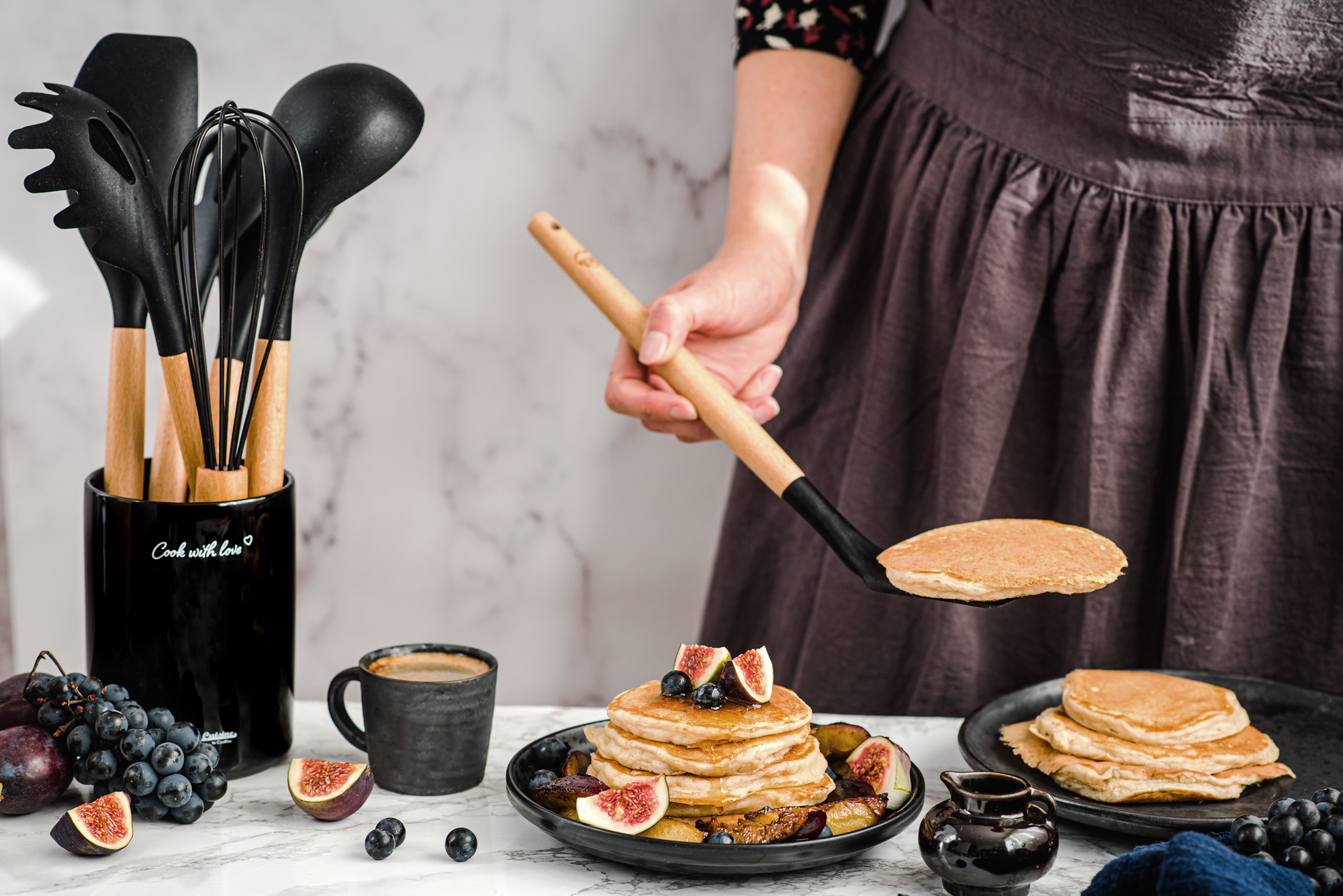 Przybory kuchenne w kolorze czarnym stojące na blacie. Obok kobieta nakładająca upieczone placki na talerz.