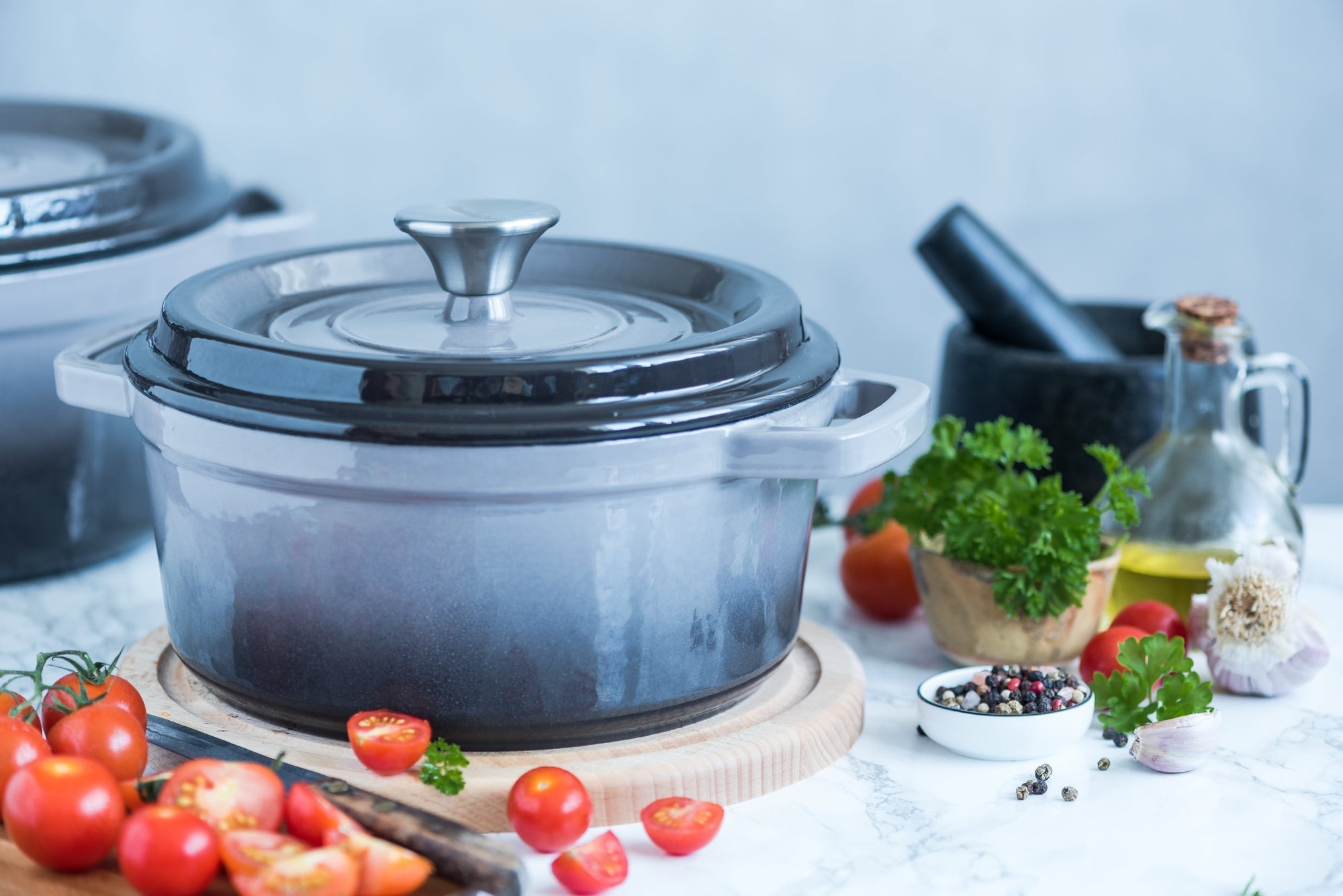 Czarny żeliwny garnek ombre z pokrywką. Obok garnka leżą pomidorki koktajlowe. Na stole stoi karafka z oliwą, moździerz i pieprz ziarnisty.