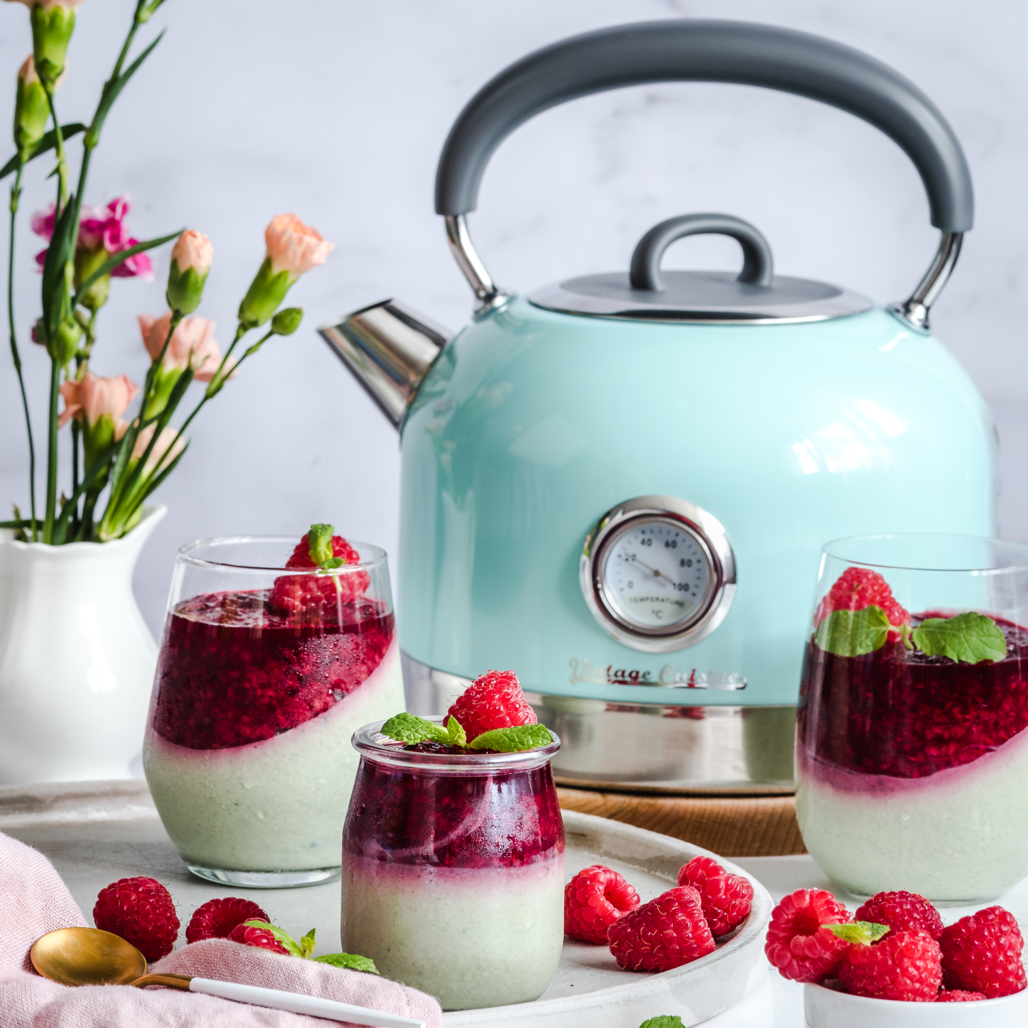 Miętowy czajnik z termometrem stojący na drewnianej podstawce na stole. Obok desery w szklankach i wazon z kwiatami.