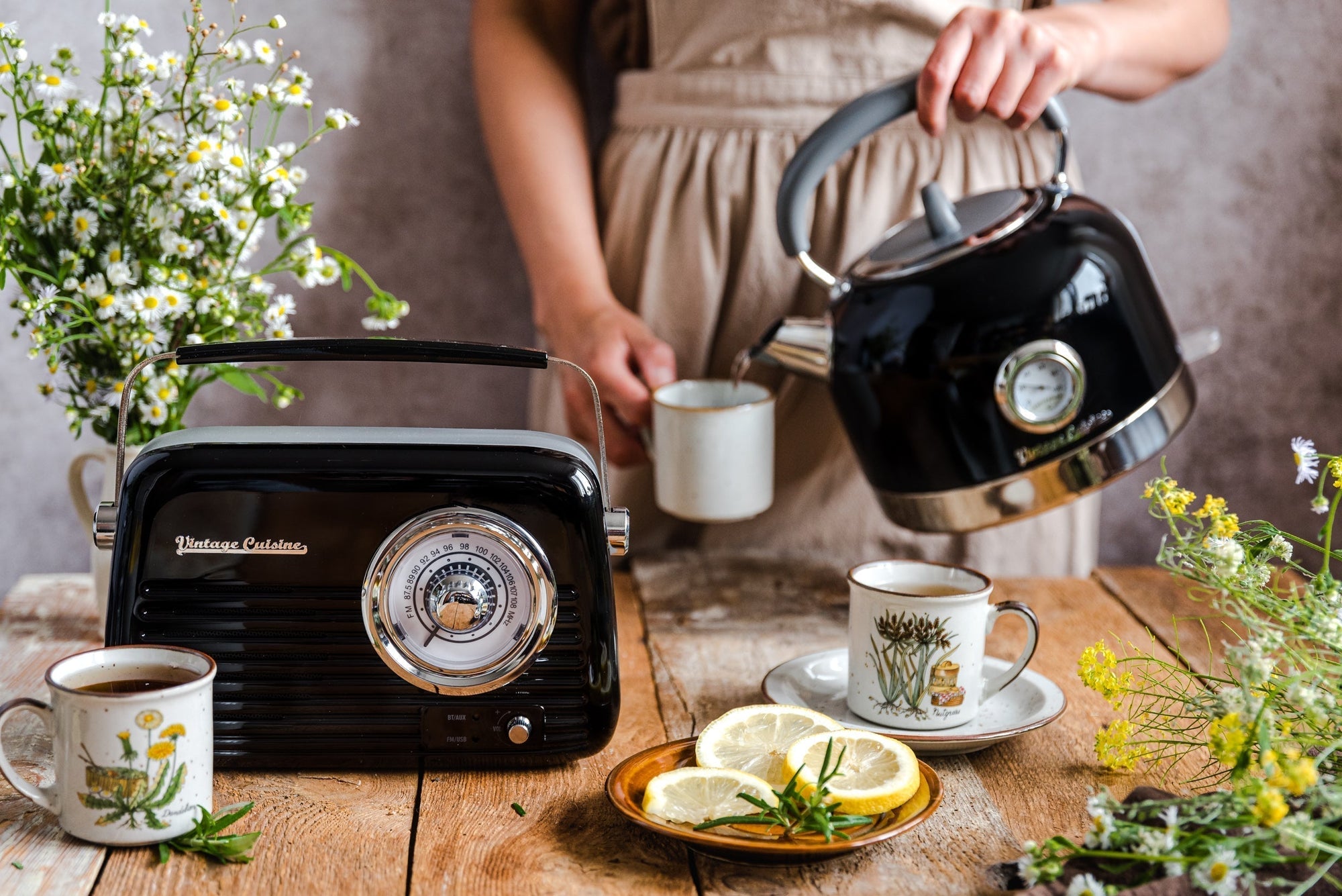 Czarne radio w stylu vintage stojące na drewnianym stole. Obok niego kubki z herbatą i plasterki cytryny na talerzu. W tle kobieta nalewa wodę z czajnika z termometrem do kubka.