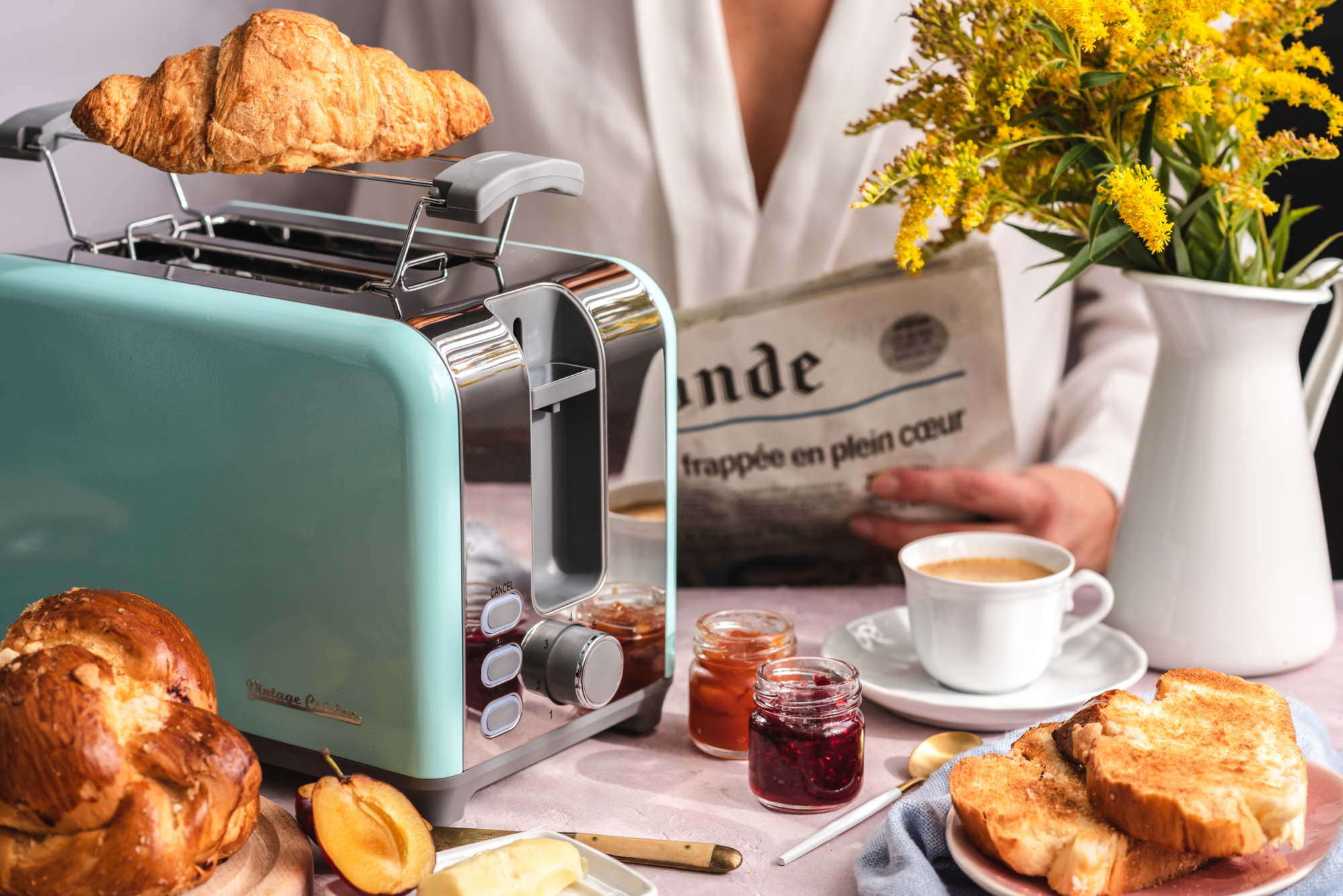 Toster z rusztem w kolorze mięty. Na ruszcie kobieta układa przekrojoną bułkę. Obok stoi talerzyk a na nim kanapka z pesto, jajkiem i pomidorem. W tle widać doniczkę z bazylią.