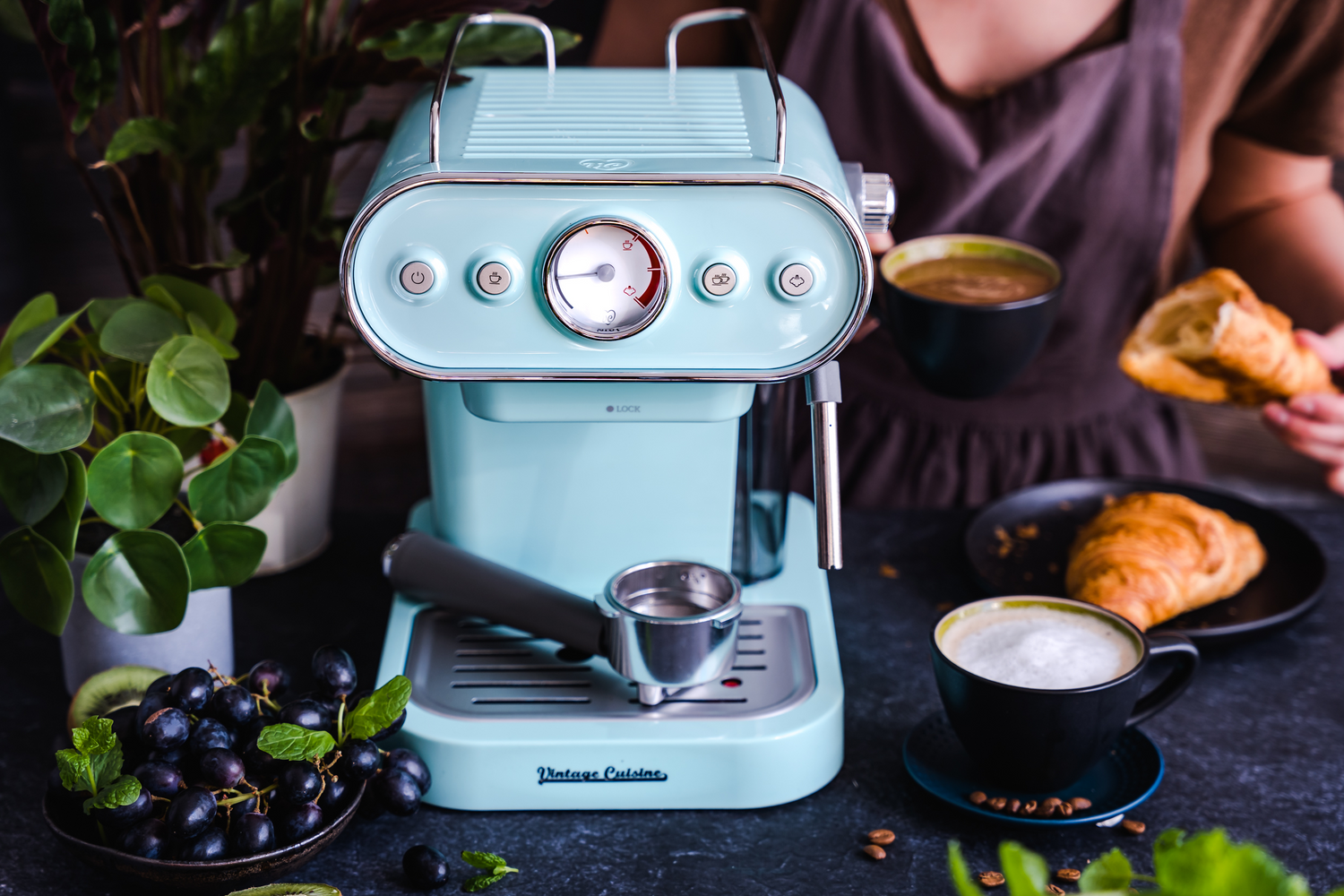 Miętowy ekspres do kawy stojący na ciemnym blacie. Obok niego filiżanka z mleczną kawą, miseczka ze śliwkami i kwiat. Za ekspresem siedzi kobieta, która trzyma kawę i croissanta.