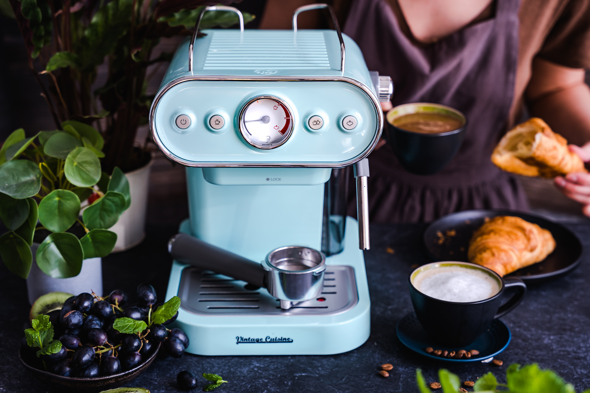Miętowy ekspres do kawy stojący na ciemnym blacie. Obok niego filiżanka z mleczną kawą, miseczka ze śliwkami i kwiat. Za ekspresem siedzi kobieta, która trzyma kawę i croissanta.