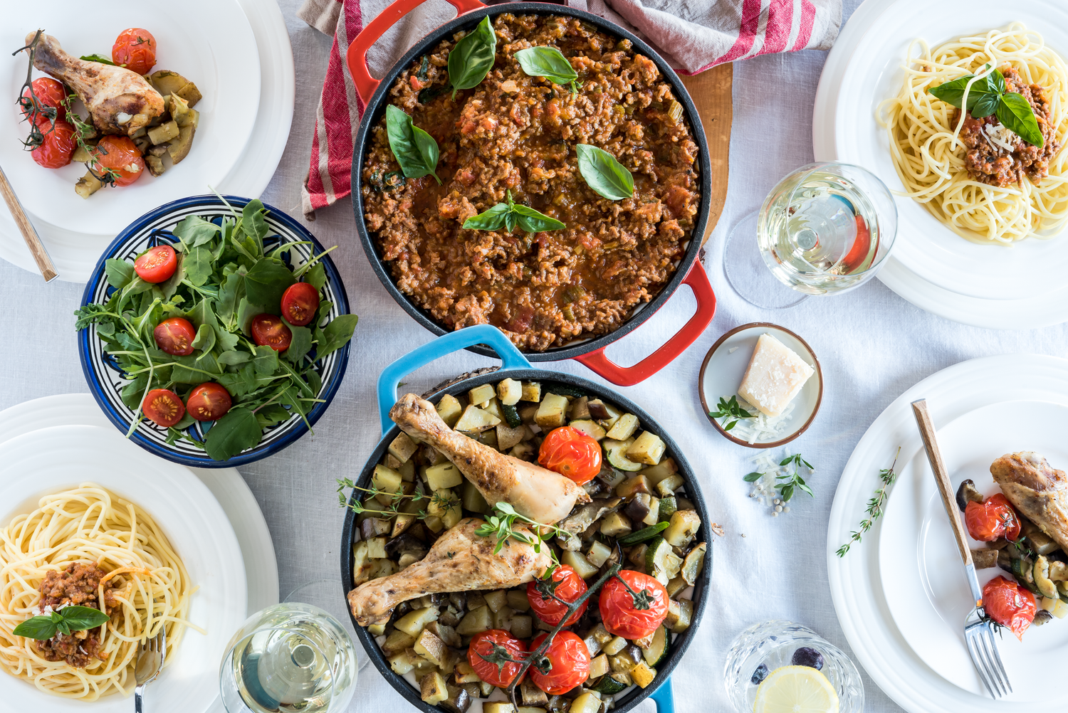 Garnki żeliwne w dwóch kolorach widziane z góry. W garnkach 2 różne potrawy. Obok talerze z nałożonymi daniami, sałatka, kieliszki z białym winem.