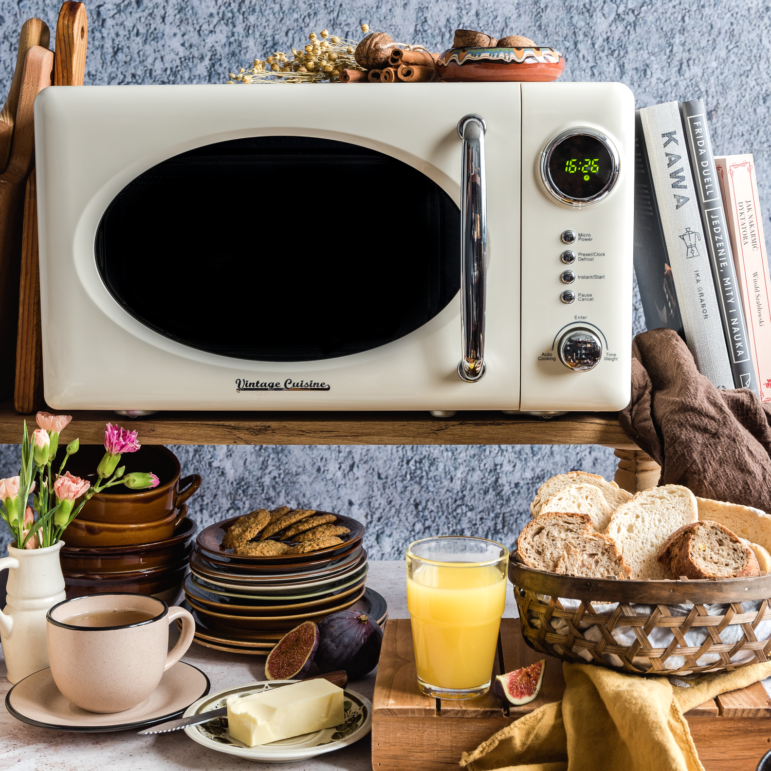 Kuchenka mikrofalowa w kolorze kremowym stojąca na półce. Obok niej książki, a pod nią talerzyki z ciastkami, kawa i sok w szklance.