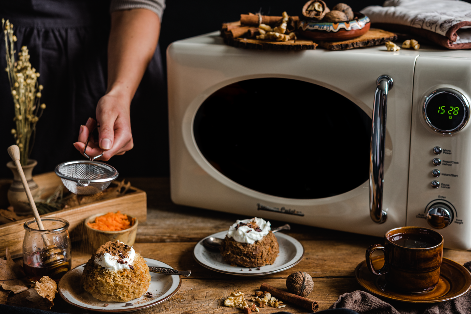 Kuchenka mikrofalowa w kolorze kremowym. Obok widoczna ręka kobiety z sitkiem. Przed kuchenką stoją talerzyki z ciastkami z bitą śmietaną oraz filiżanka z napojem.