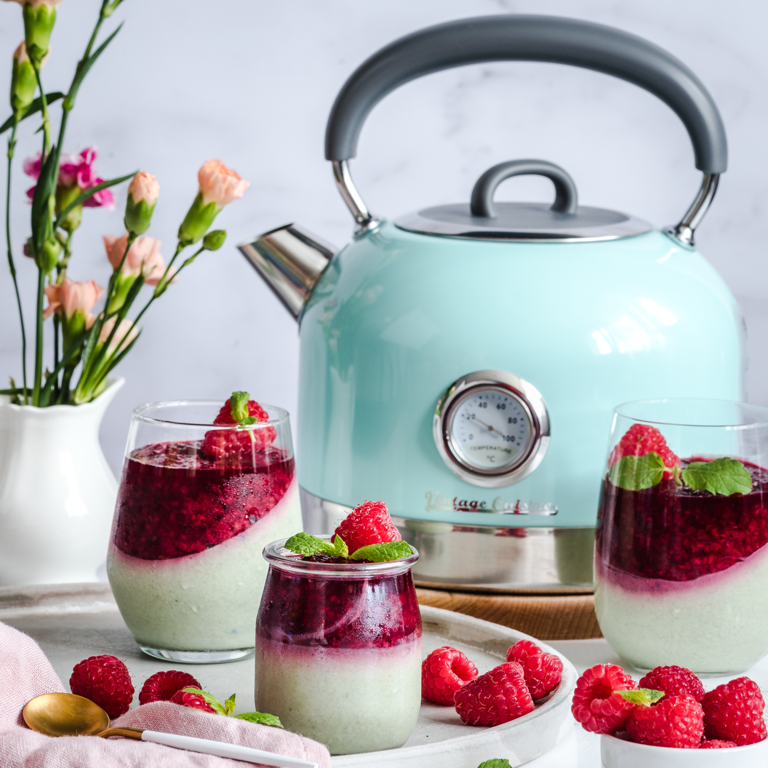 Miętowy czajnik z termometrem stojący na drewnianej podstawce na stole. Obok desery w szklankach i wazon z kwiatami.