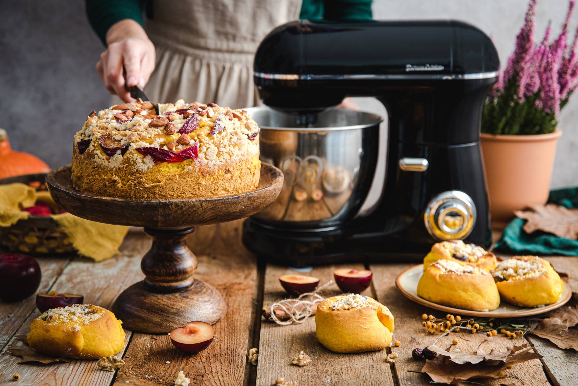 Mikser planetarny w kolorze czarnym. Przed mikserem stoi taca z ciastem. Widoczna kobieca ręka krojąca ciasto. Na talerzu słodkie bułki i jesienna dekoracja