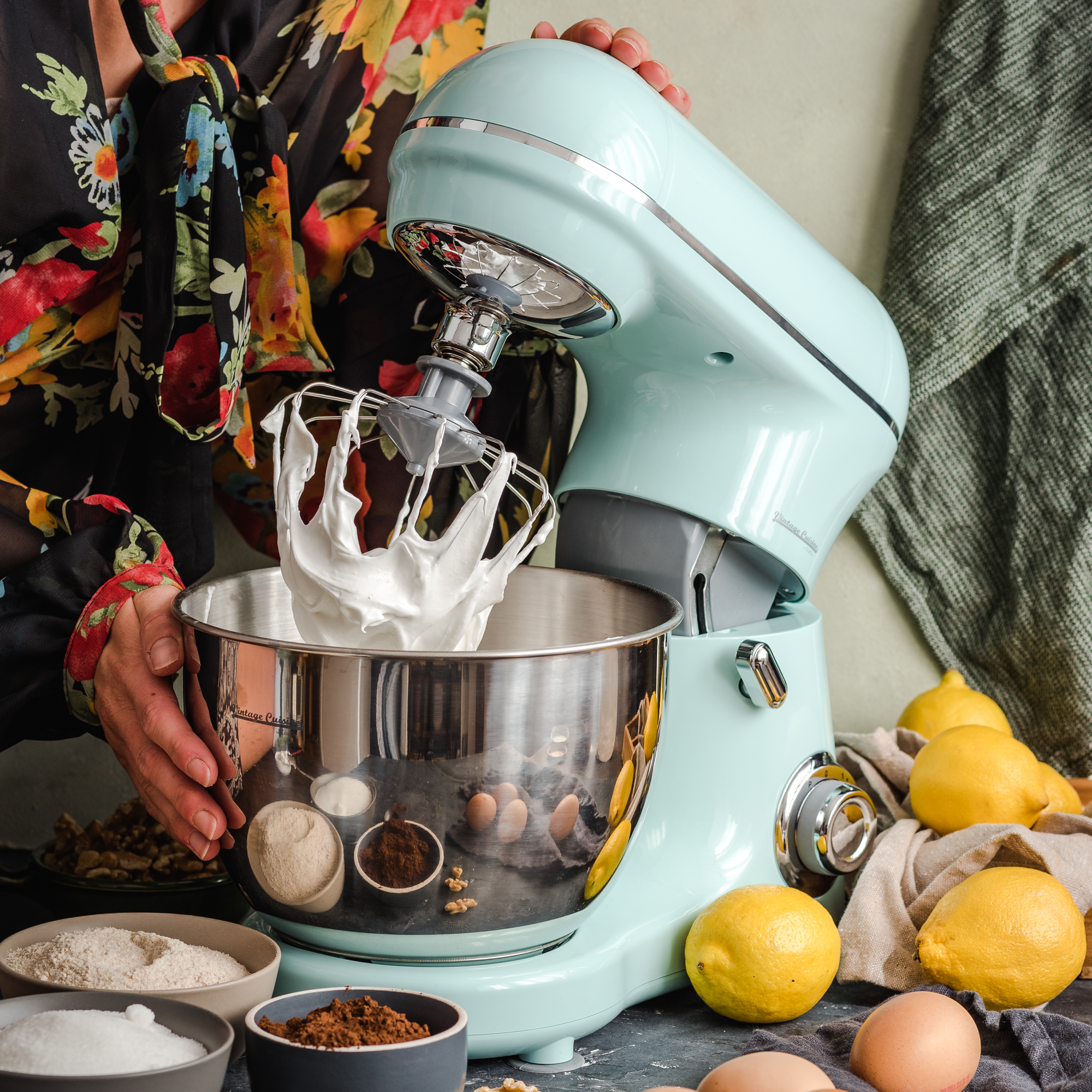 Kobieta obsługująca mikser planetarny w kolorze miętowych. Na zdjęciu widoczna jest końcówka do ubijania białek. Obok miksera leżą jajka, cytryny, stoją też miseczki z cukrem, kakao i mąką.