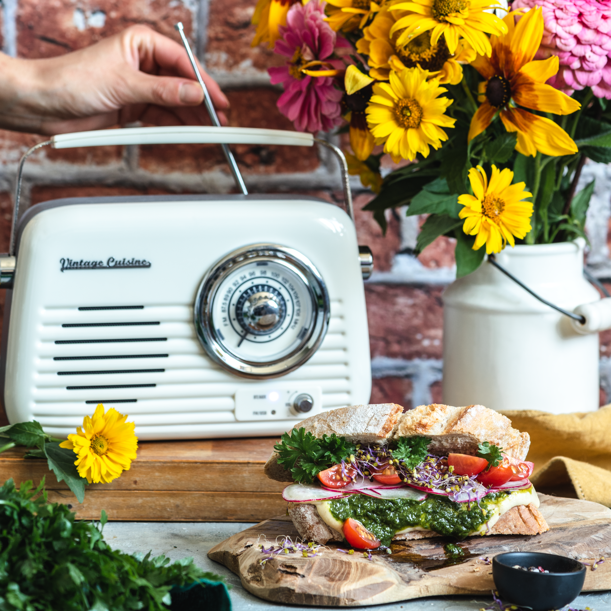 Radio w kolorze kremowym stojące na drewnianej desce. Przed radiem znajduje się kanapka z pesto i dodatkami. Obok radia stoi wazon z wiosennymi kwiatami. Kobieca ręka wyciąga antenę z radia.