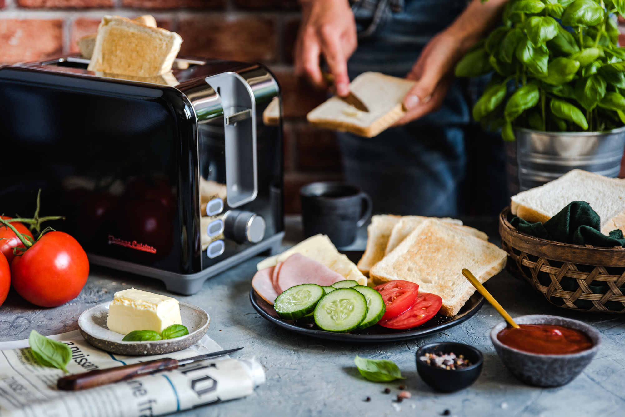 Czarny toster w stylu retro z 2 tostami. Obok widoczne dłonie osoby smarującej tost masłem. Obok tostera stoi talerz z tostami, szynką, serem, ogórkami i pomidorami. Przed tosterem leży gazeta, a na niej stroi talerzyk z masłem i leży nożyk.