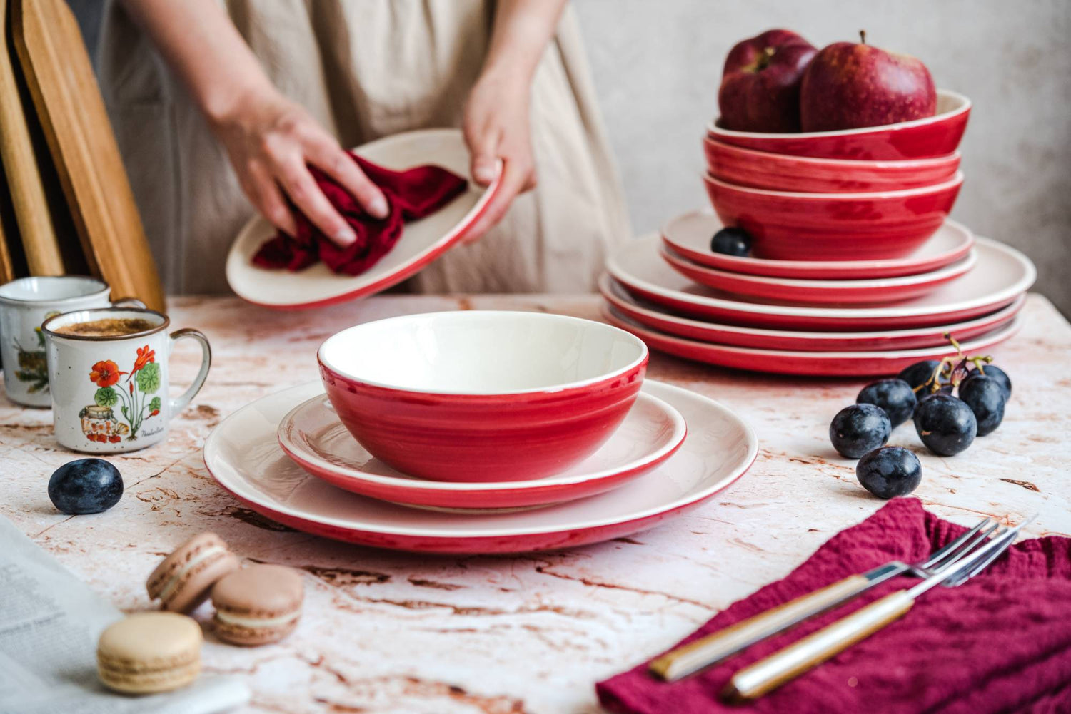 Czerwona zastawa stołowa ceramiczna stojąca na stole. Kobieta wyciera ściereczką talerz ceramiczny. Na stole stoją 2 kubki z napojem, leżą winogrona i ciastka z nadzieniem.